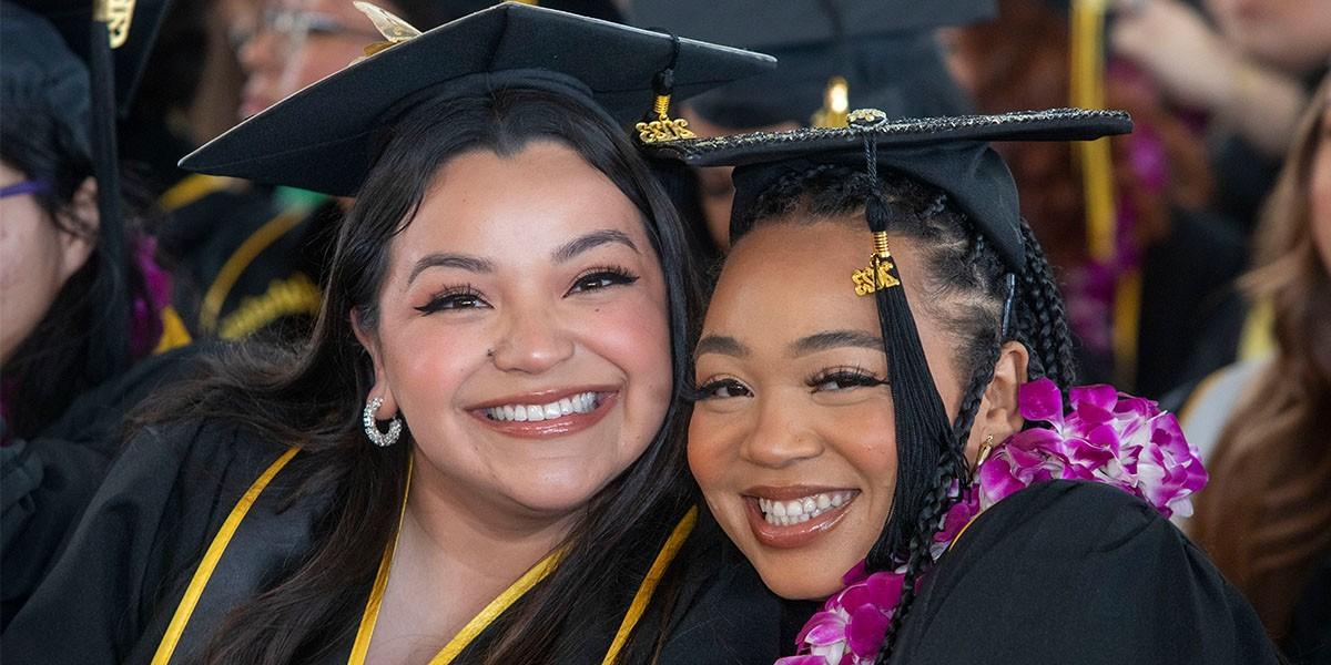 Two graduates cheek to cheek at commencement ceremony wearing graduation robe